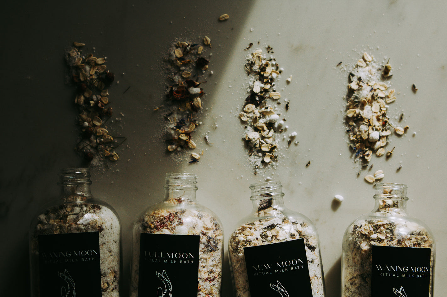Four clear bottles lay on their sides with a mix of botanicals, salt, and oatmeal spilling out.