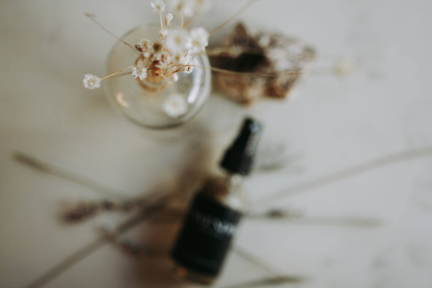an out of focus rose spray bottle lays behind a jar of white flowers