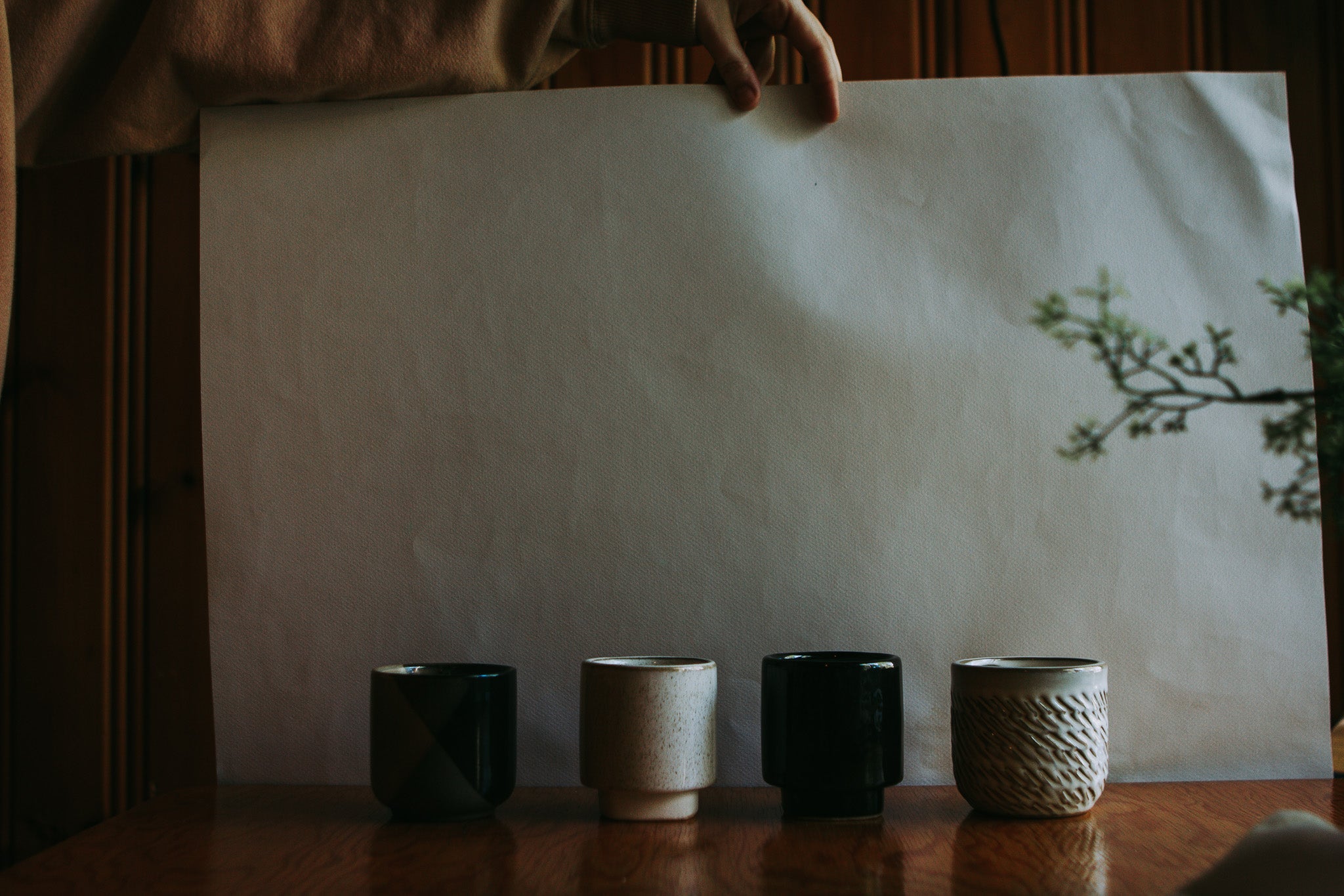 Four ceramic cups in black and beige tones lined up on a wooden table, with a hand holding a large blank sheet of paper behind them in a dimly lit setting.