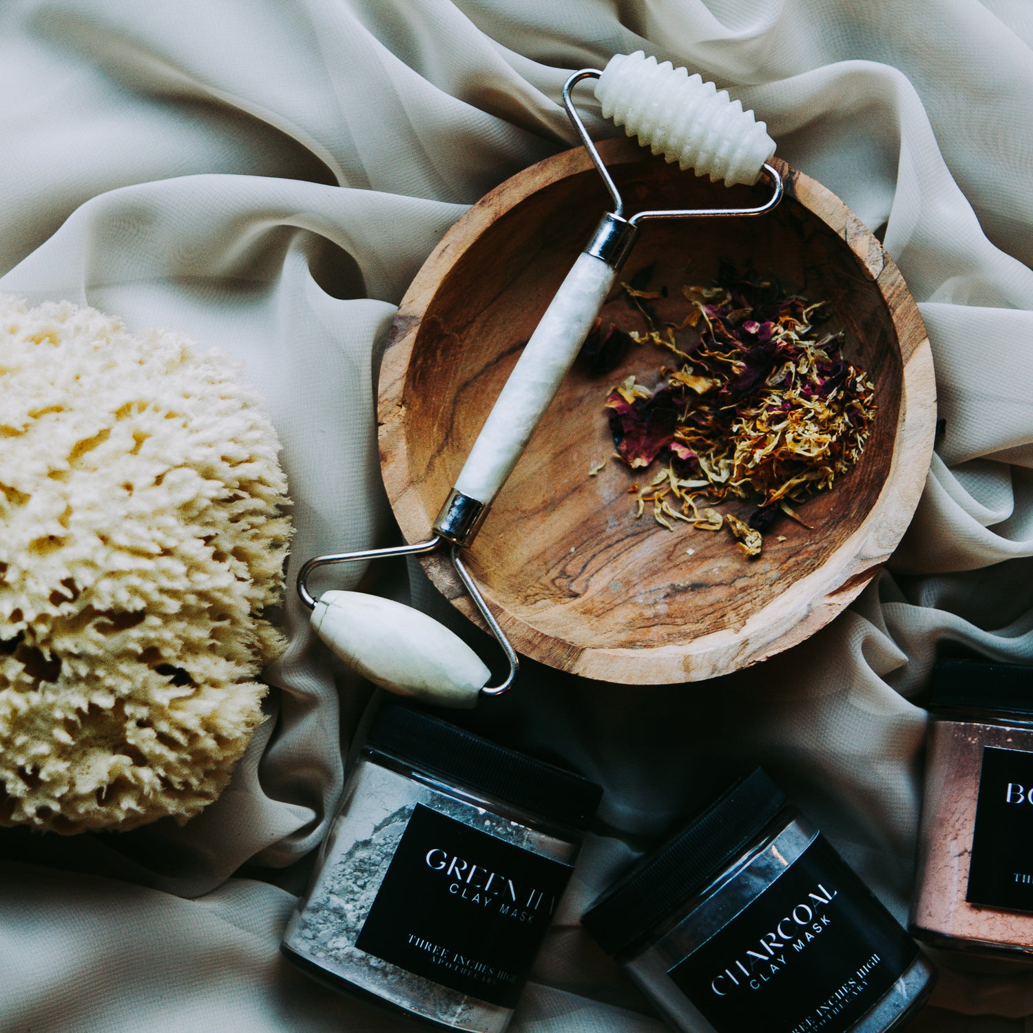 jade face roller sits atop a wooden bowl with dried botanicals. nearby Three Inches High clay face masks lay on thier sides