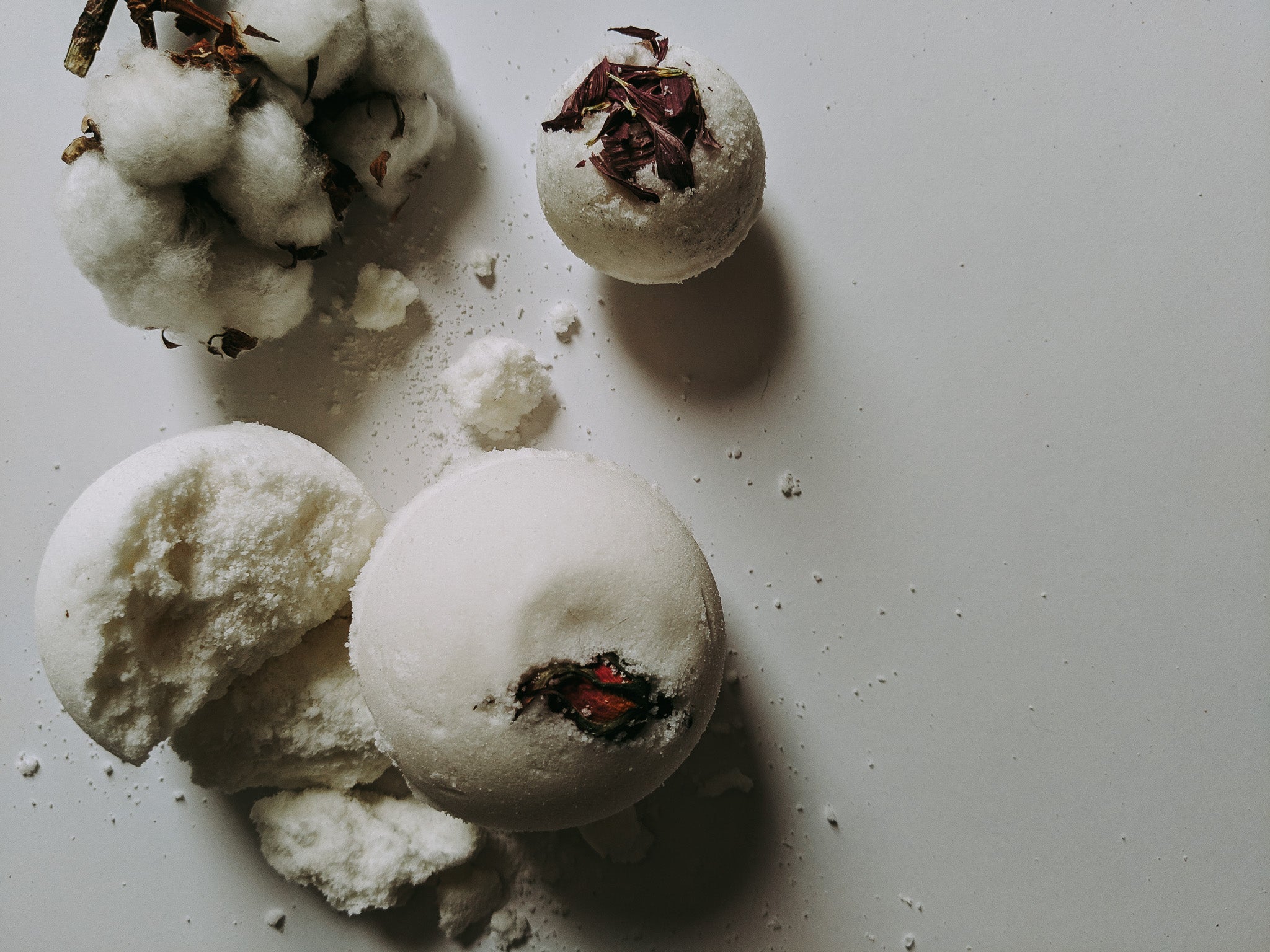 Three white bath bombs with dried botanical pieces on top, arranged on a light gray surface with some powder scattered around.