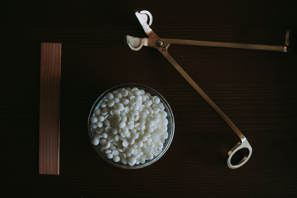 A glass bowl filled with small white wax melts or beads, placed on a dark wooden surface alongside a brass temperature gun and a wooden thermometer strip.