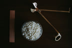 A glass bowl filled with small white wax melts or beads, placed on a dark wooden surface alongside a brass temperature gun and a wooden thermometer strip.