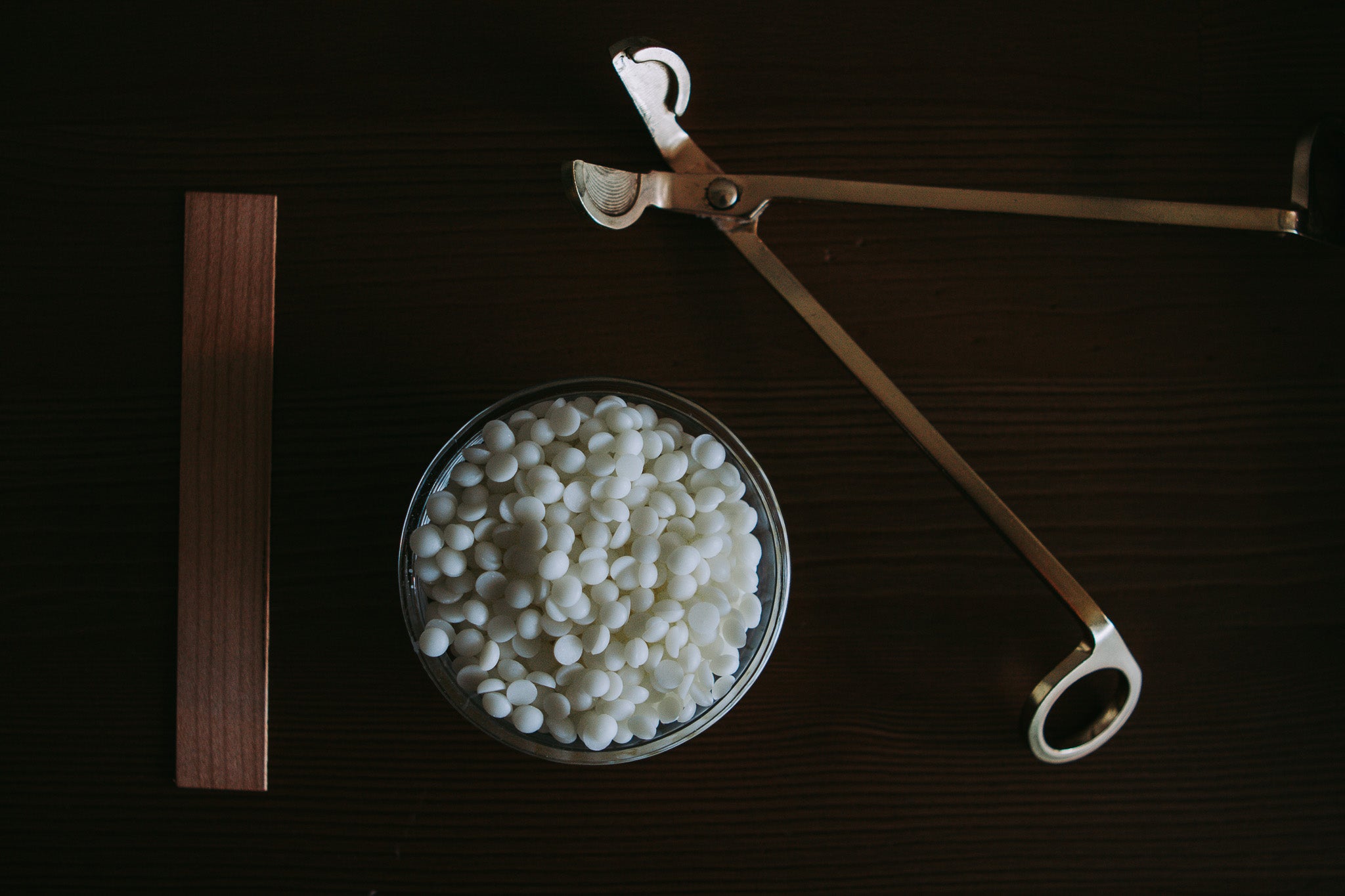 A glass bowl filled with small white wax melts or beads, placed on a dark wooden surface alongside a brass temperature gun and a wooden thermometer strip.