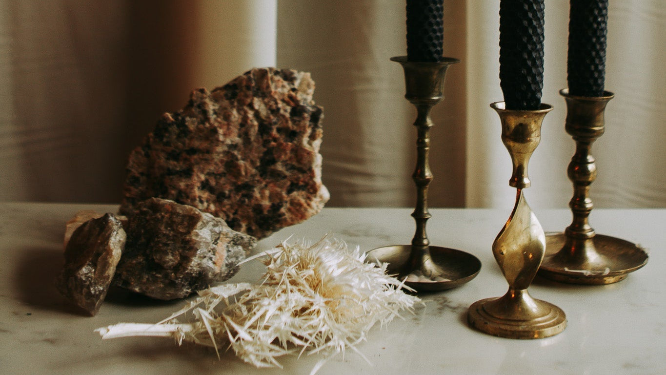 Three black taper candles in brass and bronze candlesticks, styled with raw rock crystals and dried white grass on a light marble surface in front of beige curtains.