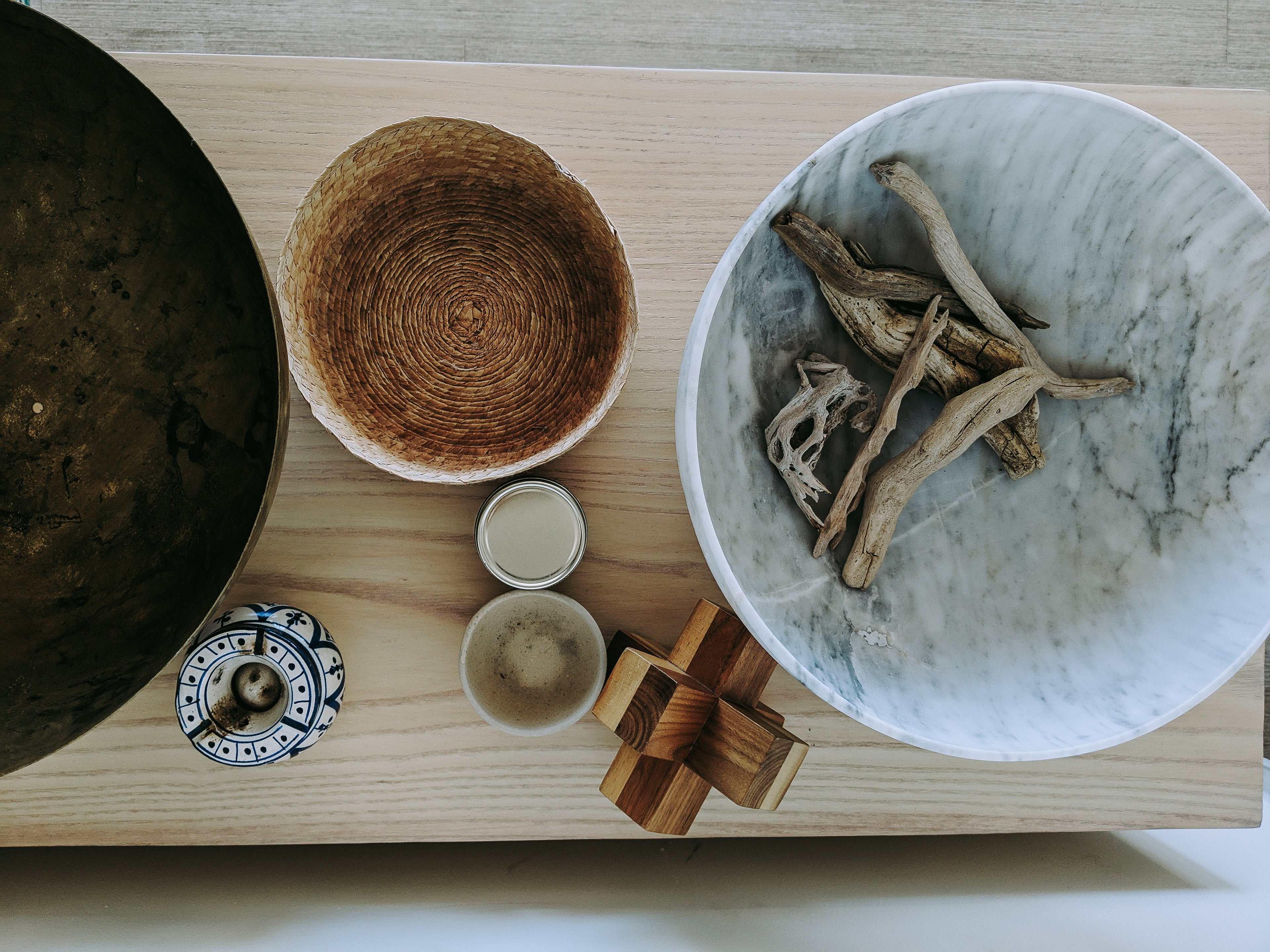 A styled flat-lay of home decor and kitchenware on a light wood surface, featuring a large white marble bowl with driftwood, a woven brown bowl, a dark metal bowl, two small ceramic cups, a blue and white patterned dish, and a wooden geometric weight.

