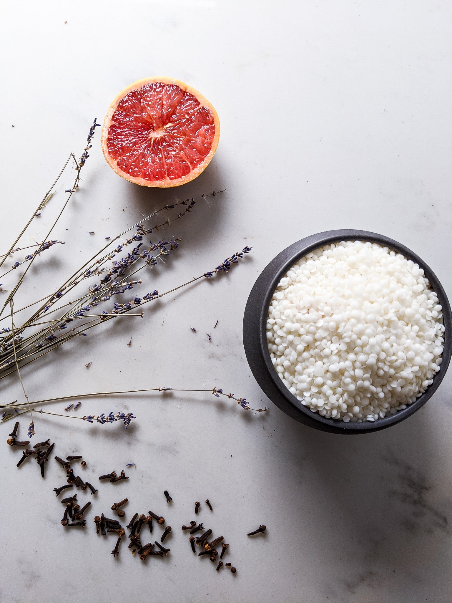 A styled flat-lay of white bath salts in a black bowl, accompanied by a halved grapefruit, dried lavender sprigs, and scattered lavender buds on a light marble surface.